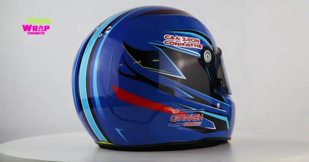 A custom-wrapped full-face racing helmet displayed on a white stand. The design features a royal blue base with dynamic speed-line graphics in cyan, black, red, and yellow-green accents. "The Carwash Company" is branded on the lower side. Finished in 3M IJ180mC-10 with 8518 Gloss laminate. Designed and installed by Vinyl Wrap Toronto.