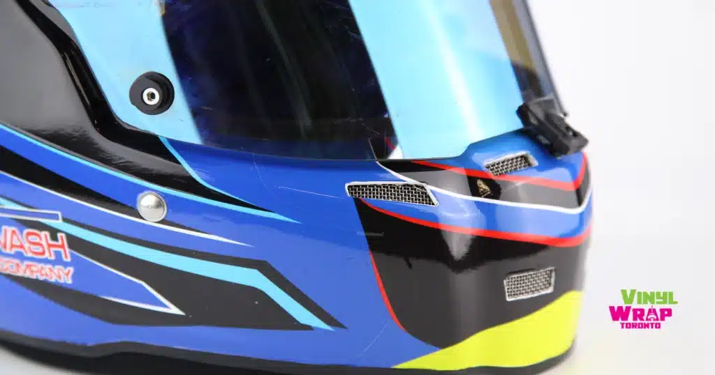 A custom-wrapped full-face racing helmet displayed on a white stand. The design features a royal blue base with dynamic speed-line graphics in cyan, black, red, and yellow-green accents. "The Carwash Company" is branded on the lower side. Finished in 3M IJ180mC-10 with 8518 Gloss laminate. Designed and installed by Vinyl Wrap Toronto.